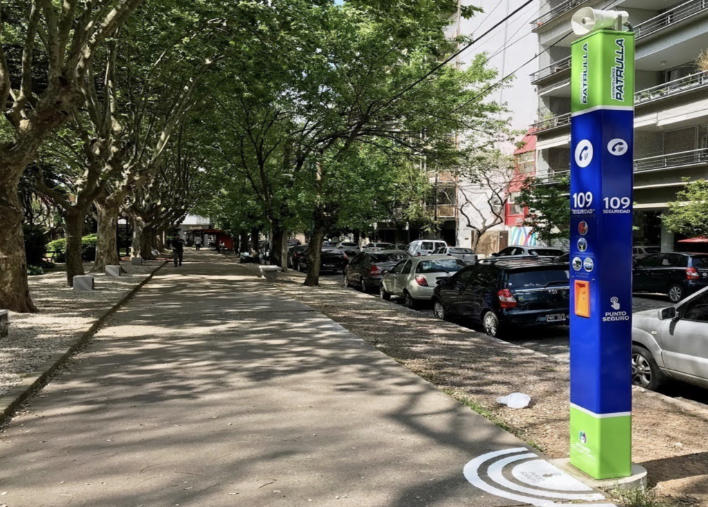 Tótem de seguridad en una calle urbana moderna, equipado con botón de pánico, cámara domótica de alta resolución, conectividad WiFi y celular, y comunicación bidireccional.